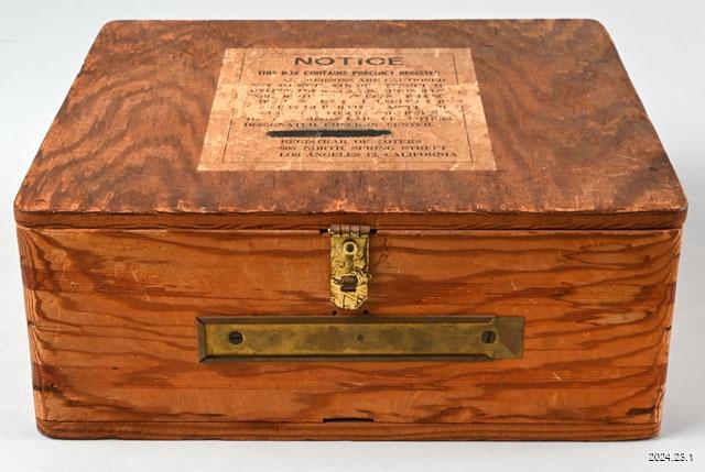 A vintage wooden box with a brass latch and a rectangular brass label holder. The top has a printed notice with text in capital letters. The wood has a textured grain and a warm, natural color.