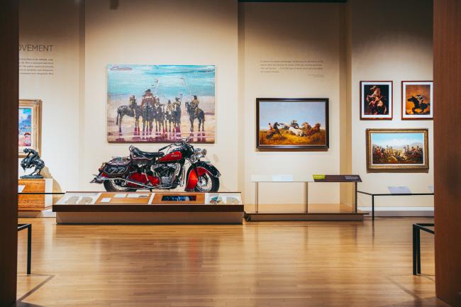 Museum gallery featuring a vintage motorcycle on display, surrounded by paintings depicting historical scenes. Warm lighting highlights artwork and wooden flooring enhances the elegant ambiance. Descriptive plaques adorn the walls.