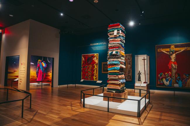 Art gallery showcasing colorful paintings and a tall sculpture made of stacked textiles. The artworks feature vibrant scenes, including religious and abstract themes. The room has wooden floors and dark walls illuminated by ceiling lights.