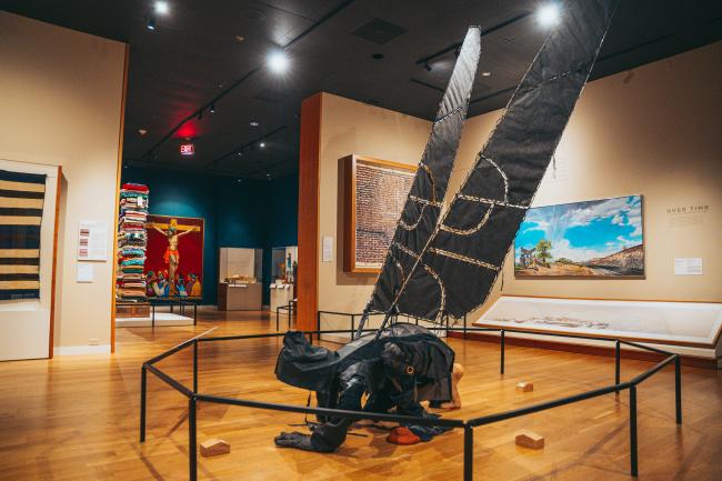 A museum exhibit featuring a sculpture of a figure with large, dark wings kneeling. The room is adorned with various artworks, including colorful paintings and textile displays. Wooden flooring and soft lighting create a warm atmosphere.