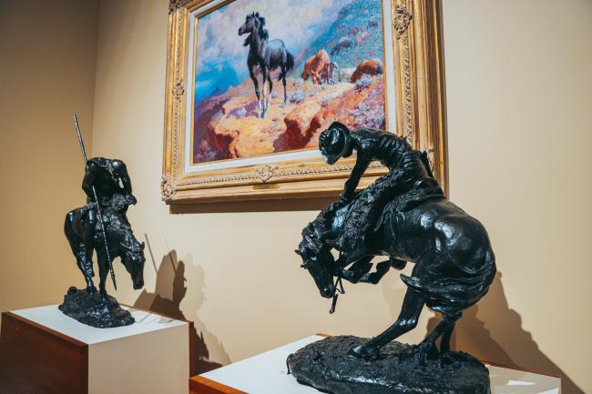 Two sculptures of horseback riders are displayed on pedestals in a museum. One sculpture is in the foreground, another slightly behind. A framed painting of a horse on a rocky terrain hangs on the wall behind them.
