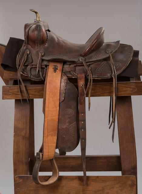 A worn brown leather Western saddle with a horn, stirrups, and saddle strings rests on a wooden saddle stand against a plain gray background.
