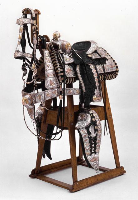 An ornate Western horse saddle with intricate silver decorations and engraved patterns sits on a wooden saddle stand. The initials "EHB" are visible on one of the saddle's protective flaps.