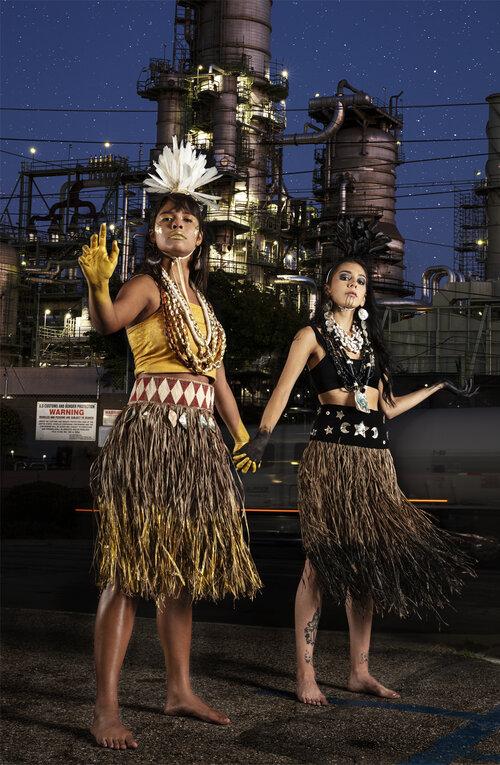 Two women wearing traditional Pacific Islander attire, with grass skirts, jewelry, and feathered headpieces, stand barefoot and hold hands in front of an industrial refinery at night under a starry sky.
