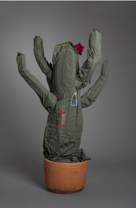A fabric sculpture resembling a cactus with arms, made from green padded material, decorated with patches and embroidery, stands in a patterned terracotta pot against a plain gray background.