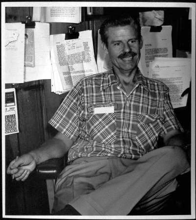 A man with a mustache, wearing a plaid short-sleeve shirt, sits smiling in an office chair. Behind him, papers and documents are clipped to a bulletin board on the wall.
