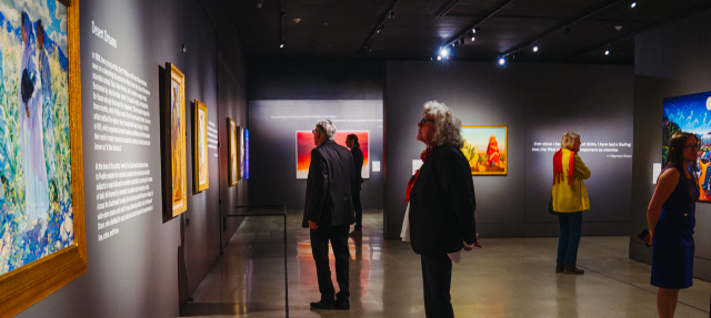 People view colorful paintings on the walls of a dimly lit art gallery. The space has polished floors, spotlights illuminating the artwork, and visitors standing and reading descriptions or admiring the art.