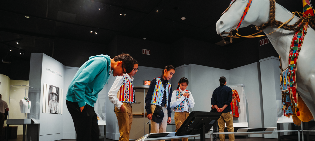 Several people observe exhibits in a museum, including a white horse statue decorated with colorful beadwork. The room has art displays and black-and-white photographs on the walls.