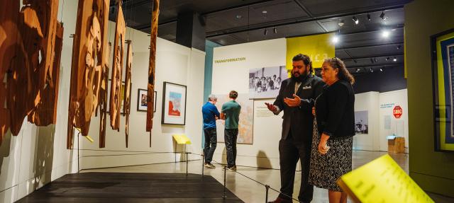 Two people stand in a museum exhibit, discussing wooden hanging art pieces. Other visitors observe displays in the background. The space is well-lit with informational signs and photos on the walls.