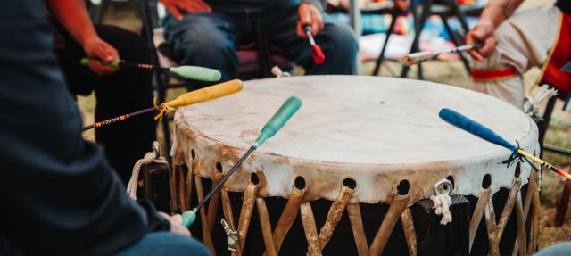 Four people sit around a large traditional drum, each holding a colored mallet and striking the drum together. The drum has a taut, laced surface and is played outdoors.