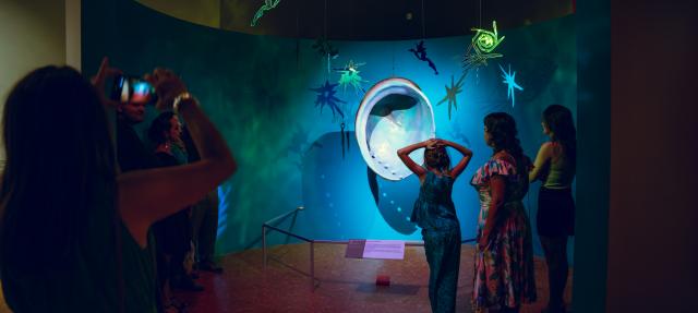 Visitors in a dimly lit museum gallery look at a large, glowing, circular shell-like sculpture suspended before a wall decorated with illuminated, hanging fairy and star shapes.