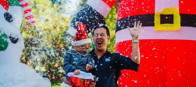 A man holding a toddler in a Santa hat while artificial snow falls in front of giant inflatable holiday decorations.