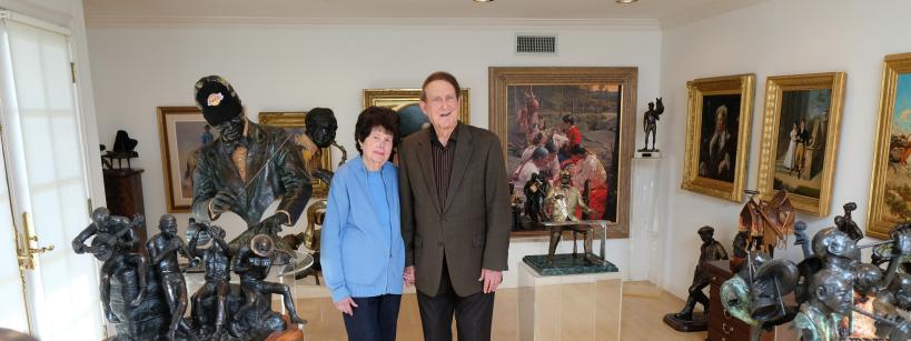 An older couple stands smiling in an art-filled room. They are surrounded by various sculptures and framed paintings on the walls. The sculptures include human figures and animals, displayed on pedestals and shelves. The room is well-lit.