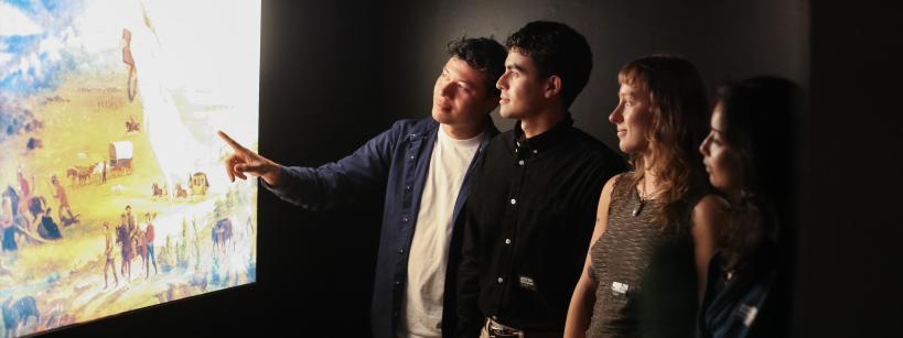 Four people stand in a dimly lit room, attentively viewing and discussing a large, illuminated painting on the wall. One person points toward the artwork, as the others look on with interest.