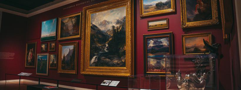 A gallery with framed paintings on red walls displays various landscapes and portraits. Glass cases exhibit silver artifacts. Wooden benches provide seating. The room is softly lit, enhancing the art's details.