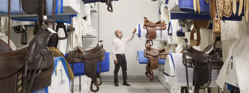 A man in a white shirt and black pants examines intricately designed saddles in a storage room. The saddles are mounted on racks, arranged on blue and white protective padding. The room has shelves and a bright atmosphere.
