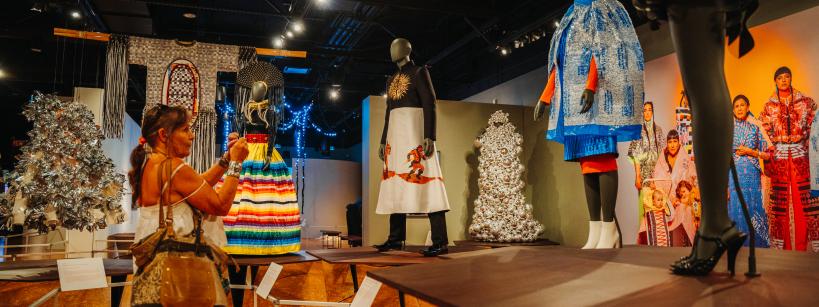 A woman takes a photo of mannequins dressed in colorful, elaborate outfits on display in a museum exhibit with dramatic lighting and various artwork in the background.