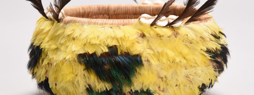 A round woven basket decorated with yellow and green feathers, brown feathers sticking up at the rim, and white shell-like ornaments; the background is plain light gray.