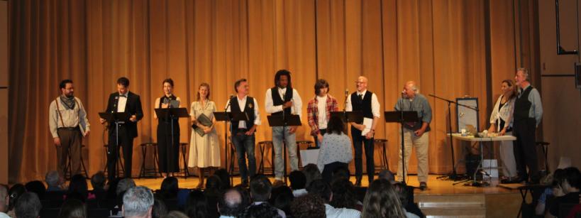 A group of people stand on a stage in front of a beige curtain, some holding scripts. An audience is seated in the foreground, watching the performers. A person stands in front of the stage, possibly directing or addressing the group.
