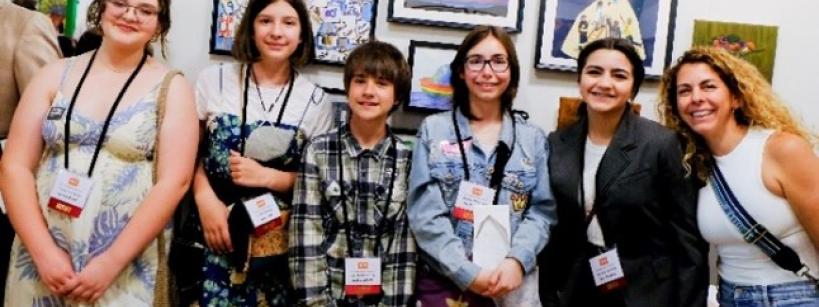 A group of six people, including five young students and one adult, stand smiling in front of an art display featuring colorful paintings and a collage reading "We the People." All wear nametags and casual to semi-formal attire.