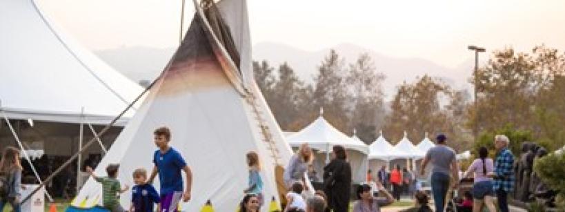 People, including families and children, gather around a large tipi and several white tents at an outdoor event on a sunny day, with trees and mountains visible in the background.