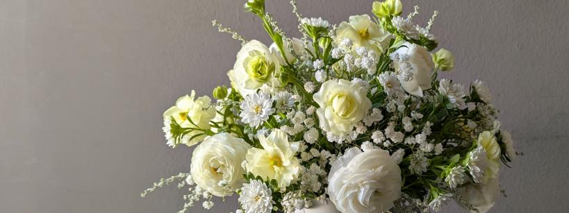 A white ceramic vase filled with white and pale yellow flowers, including roses and daisies, sits on a black table against a plain gray background.