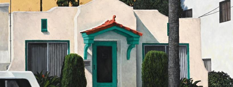 A small white stucco house with a red tile roof and green trim on the door and windows, surrounded by bushes and palm trees, with part of a white car visible on the left.
