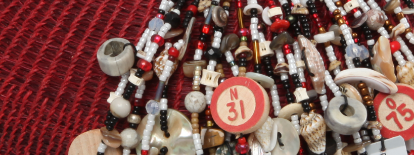 A close-up of a beaded necklace with assorted beads, shells, and numbered bingo chips, all resting on a textured red woven fabric background.