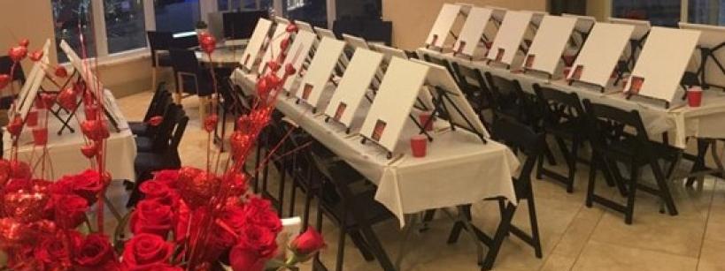 A room is set up for a painting class with rows of easels and blank canvases on tables, each with a red cup. In the foreground, a bouquet of red roses adds a decorative touch. Large windows are visible in the background.