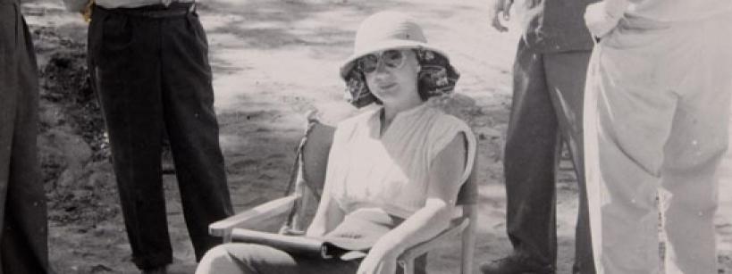 A woman wearing a light-colored hat and dress sits on a chair outdoors, holding an open book. Three men stand around her, their faces out of frame. The scene appears to be from an earlier era.