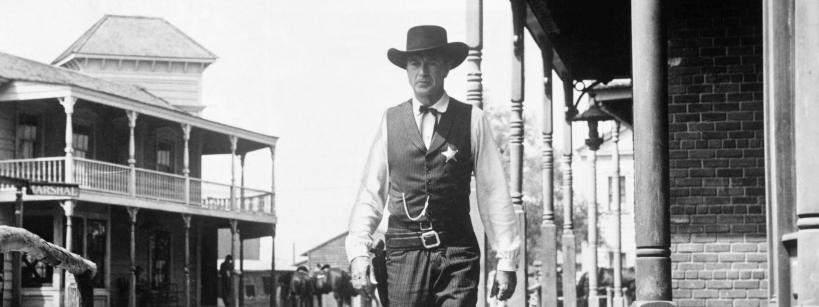 A sheriff wearing a vest, cowboy hat, and star badge walks confidently down a deserted street in a classic Western town with wooden buildings and porches in the background.