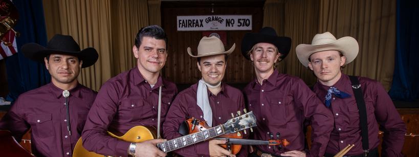 Five men in matching maroon shirts and cowboy hats pose on stage with musical instruments, including a guitar, upright bass, violin, and drumsticks. A sign reading “Fairfax Grange No 570” hangs in the background.