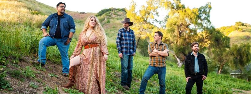 Five people stand on a grassy hillside, dressed in casual and country-style clothing. The woman in the center wears a patterned dress and boots, with trees and hills in the background under a clear sky.