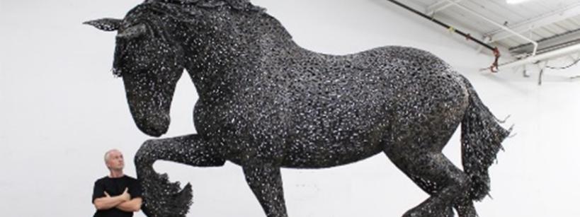 A man stands with arms crossed next to a large, detailed horse sculpture made from metal pieces, displayed indoors on a wheeled platform in a white-walled room with a high ceiling.