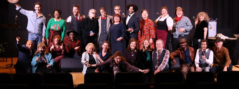 A group of around 25 people pose together on a stage, some standing and some sitting, in front of a black curtain. Several are smiling or gesturing enthusiastically, and the mood appears celebratory.