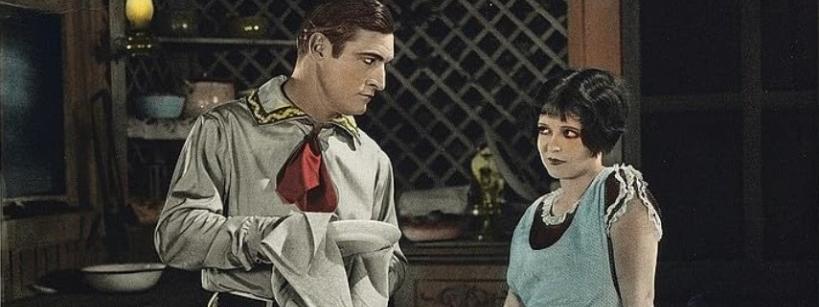 A man in a western-style shirt and red neckerchief dries a dish while looking at a woman in a blue dress, who gazes back with a skeptical expression. They stand in a rustic kitchen with shelves and dishes behind them.