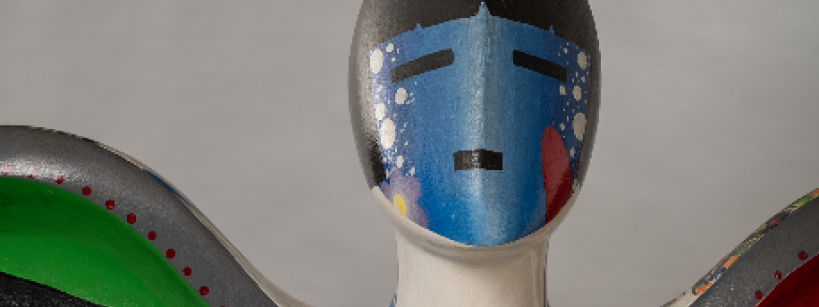 A close-up of a colorful abstract sculpture of a human figure with a smooth, painted face featuring blue and black geometric shapes, white dots, and bold green, red, and blue patterns on its shoulders.