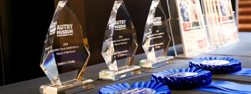 A row of glass awards labeled “Autry Museum” and 2023 sit on a black table alongside several blue ribbon rosettes, and framed certificates in the background.