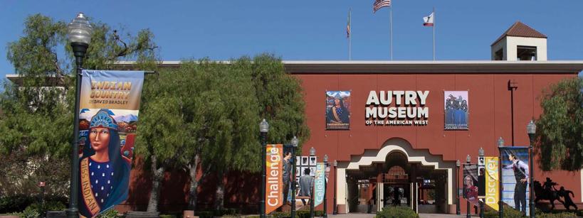 Entrance to the Autry Museum of the American West with banners reading "Indian Country," "Challenge," and "Discover," potted plants, and two flags flying above the red brick building.