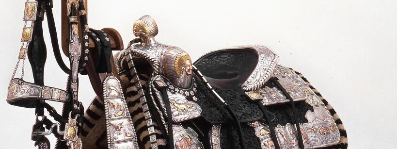 An ornate, Western-style saddle with detailed silver and gold embellishments, intricate leather tooling, and decorative straps, displayed against a plain white background.