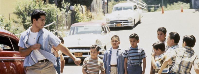 A young man poses in a fighting stance near a red car on a street, facing a group of boys who watch him. The boys appear curious or excited, and old cars are parked in the background.