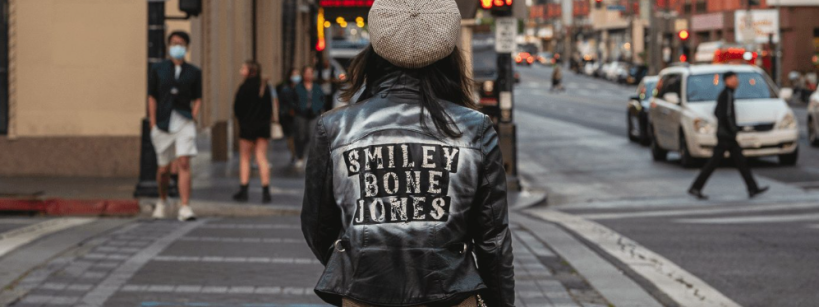 A person wearing a leather jacket with “SMILEY BONE JONES” on the back stands at a city street corner, facing away from the camera. Cars and pedestrians are visible in the urban background.